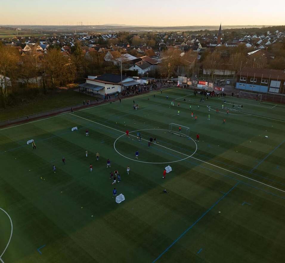 Luftaufnahme eines Fußballfeldes mit Spielern, Toren und Zuschauern sowie Häusern im Hintergrund.